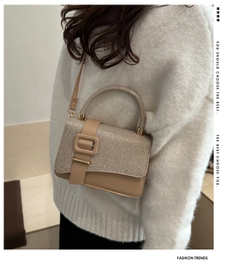 Détail du rabat pailleté et de la boucle carrée du sac beige porté sur un pull blanc