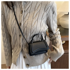 Detail of a micro black crossbody bag with wave-edged flap on a beige knit cardigan