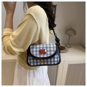 Sac à carreaux bleu et blanc avec écusson pomme rouge porté à l'épaule dans un décor doux