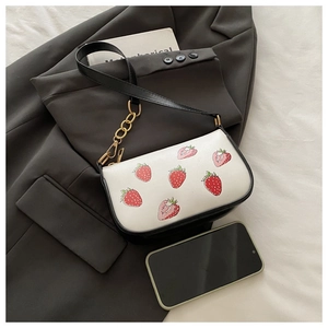 Detail der Schultertasche mit Erdbeerdruck, schwarzem Gurt und Goldkette neben einem Handy