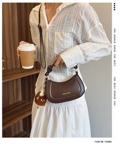 Detail of a brown crescent saddle bag with a plush charm held by a woman in a casual style