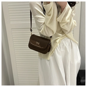 Brown saddle crossbody bag with gold bow clasp on beige sweater background