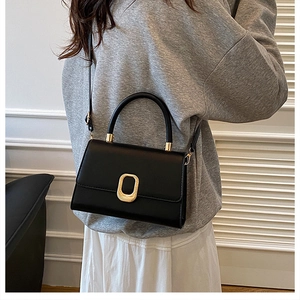 Detail of a black matte leather handbag with a textured gold oval clasp on a grey outfit