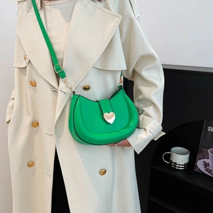 Green crescent saddle bag with gold heart clasp and adjustable strap on a beige coat