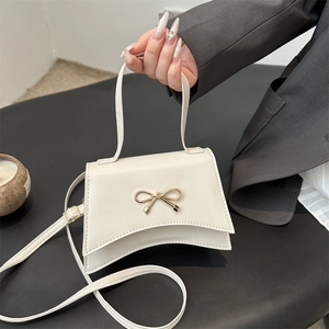 Sac à main élégant crème avec nœud doré et base incurvée porté par une main de femme