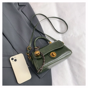 Flat lay of green leather satchel with golden clasp and bell accessory on grey blazer