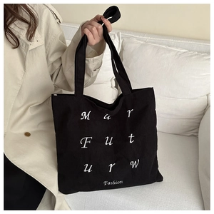 Detail of black corduroy tote bag featuring white grid letter embroidery on a white sofa