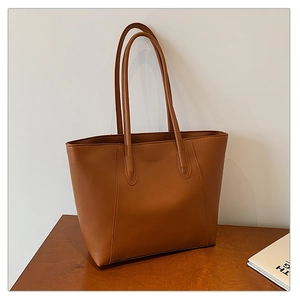 Brown leather tote bag standing on a wooden table with book, showcasing its large capacity