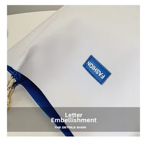 Detail of white tote bag with blue trim, fashion logo patch, and gold metal hardware