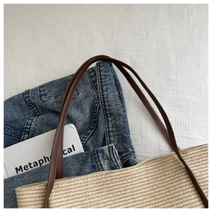 Flat lay of a woven straw tote bag with brown straps and denim jacket on a white fabric