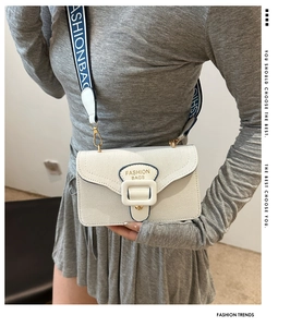 Detail of white square bag with FASHIONBAGS lettered strap and gold buckle accent