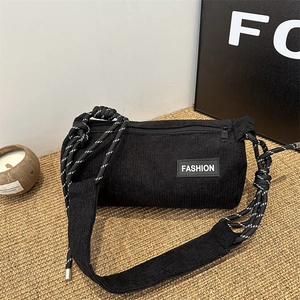 Minimalist black corduroy cylinder crossbody bag with woven rope strap on a straw mat