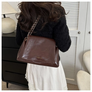 Brown glossy leather tote bag with metal chain strap worn on shoulder in a modern setting