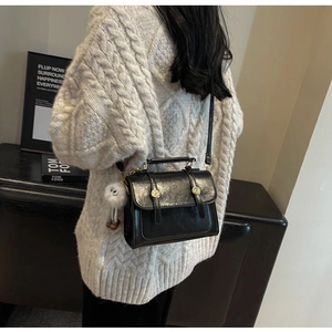Detail of a black faux leather crossbody bag with gold buckles and plush charm on white sweater