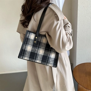 Large plaid tweed tote bag with black leather straps worn by a model in a warm interior