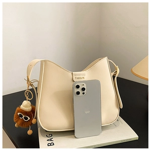 Detail of cream saddle bag with fashion label, plush charm, and phone for size scale