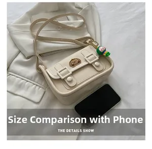 Size comparison of beige crossbody bag with embroidered bear and doll charm next to phone