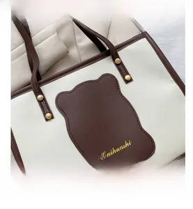 Detail of a beige and coffee tote bag with a bear-shaped patch and gold rivets