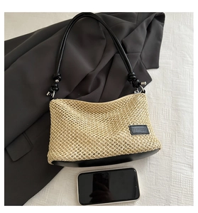 Detail view of woven beige shoulder bag with black strap and leather logo tag beside a phone