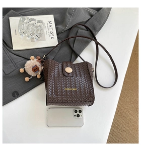 Détail du sac seau marron foncé à texture tressée avec pompon sur un fond de blazer gris