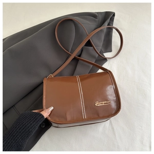 Détail du sac besace marron avec coutures blanches et logo doré sur fond gris