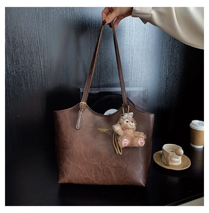 Detail of dark brown leather tote bag with gold logo and plush bear charm on wooden background