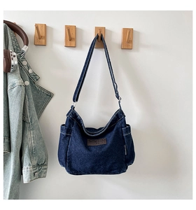 Detail of dark blue denim shoulder bag with brown leather brand patch on white wall