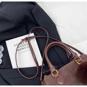 Détail du sac Boston marron foncé avec pompon posé sur un blazer noir et un livre