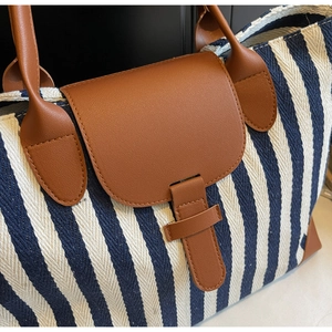 Detail of blue striped canvas tote bag showing the brown leather flap and metal buckle closure