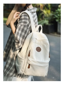 Detail of white corduroy backpack with leather logo patch worn by model over plaid shirt