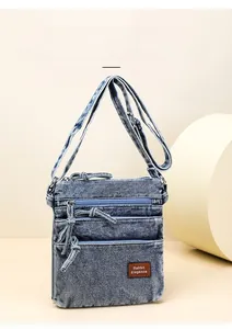 Detail of blue washed denim crossbody bag featuring multi-zipper layers and leather logo patch