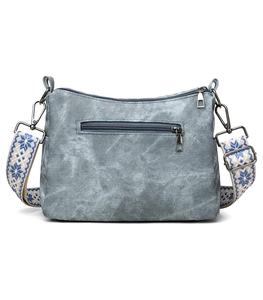 Detail of grey blue denim crossbody bag featuring a patterned wide woven strap and zippers