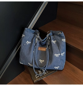 Detail of large denim tote with bow print, chain strap, and brown leather logo tag