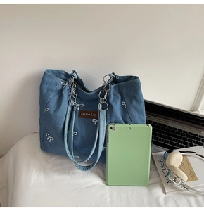Blue denim tote bag with white bow embroidery and chain strap detail with tablet for scale