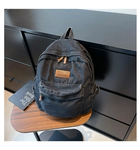 Detail of gray denim backpack showing leather logo tag and front zipper pocket on wood