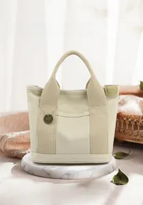 Detail of canvas tote bag with metal M logo tag and reinforced stitching on marble stand