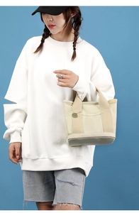 Detail of a beige canvas tote bag featuring an outer pocket and metallic M charm