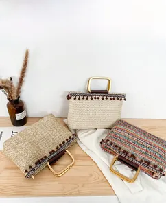 Straw tote bag detail showing colorful weave, pom-pom trim and golden metal handles