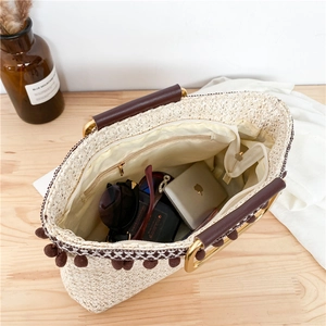 Straw tote bag with brown leather handles and decorative pom-poms on a wooden table