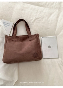 Detailed view of dark brown suede tote bag with round logo patch on white bedding