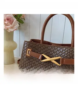 Detail view of woven brown handbag featuring a gold X-shaped hardware buckle on the front