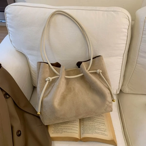 Beige suede tote bag with knotted drawstring and gold accents on a white sofa