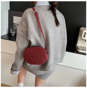 Burgundy suede oval crossbody bag shown on model in a modern interior setting
