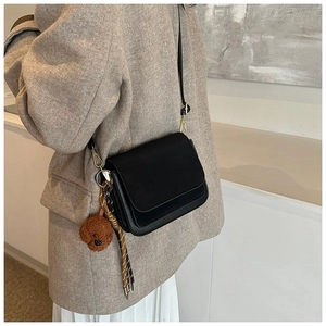 Detail of a matte black square crossbody bag featuring a cute teddy bear charm and woven cord