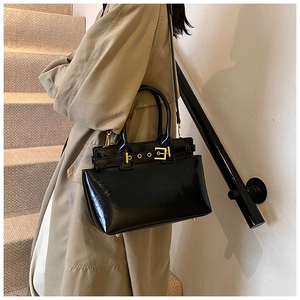Side view of black glossy patent leather handbag with gold buckle on beige coat