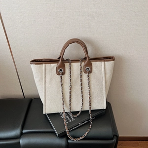 Minimalist canvas tote bag with brown leather trim and chain strap on a black bench