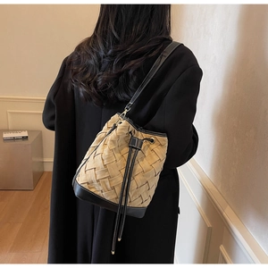 Detail of a beige woven bucket bag with black leather trim worn on shoulder by a model
