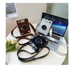 Three creative camera-shaped crossbody bags in brown, silver, and black on a white table