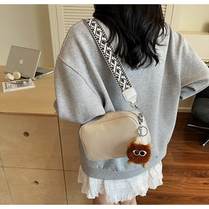 Beige crossbody square bag with jacquard strap and plush monster pendant on desk