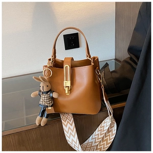 Detail of brown leather bucket bag with wide strap and rabbit plush pendant on wood table